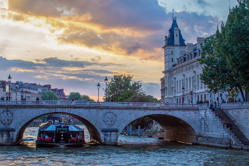 Orange sunbeams of  sunset in Parisian sky and wealthy tourists sit in cafes on a pleasure boat on the Seine and build next to Notre Dame. Also people on the waterfront. Purple-blooming chestnut tree. Unrecognizable persons in distance