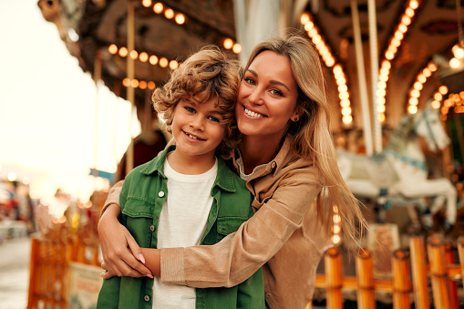 A handsome boy blonde child with a curly hairstyle together with his mother came to the amusement park to have fun and ride on the carousels and swings.