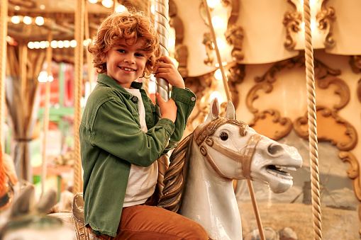 Caucasian curly-haired blonde boy ride a vintage horse carousel in the evening at an amusement park on a weekend.