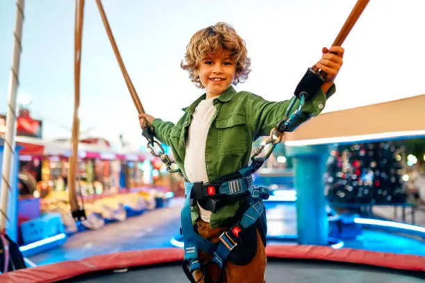 A Caucasian boy child jumping on a trampoline with insurance elastic bands in an amusement park and carousels.