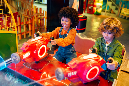 An African-American girl child with an Afro hairstyle and a cute Caucasian boy sitting in an amusement car ride playing a computer game racing in an amusement park in the evening laughing merrily and relaxing together.
