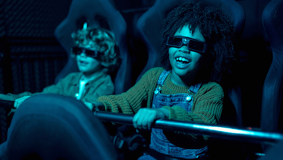 An African-American girl and a cute Caucasian boy sitting in a virtual cinema in glasses on moving rotating chairs and emotionally watching a movie in an amusement park.