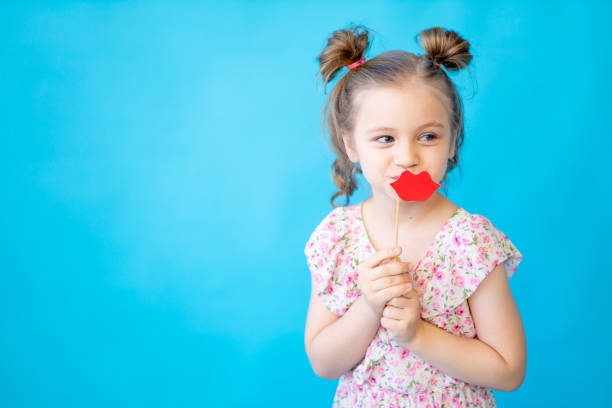 istock a little child girl on a blue isolated studio background with a paper accessory on a stick with her lips is having fun and fooling around. Space for copying text. Cute kid 1472340549