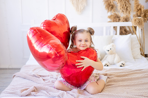 a little child girl at home in a calm natural interior with red balloons in the form of a heart is having fun and fooling around. Valentine's Day concept