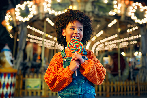 A cute African-American girl with an Afro hairstyle eats a colorful lollipop standing against the background of a carousel with horses in the evening at an amusement park or circus.