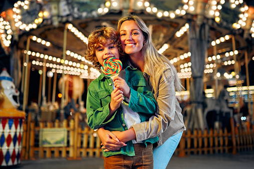 A cute caucasian boy with blonde curly hair with his mother eating a colorful lollipop standing against the background of a carousel with horses in the evening at an amusement park or circus.