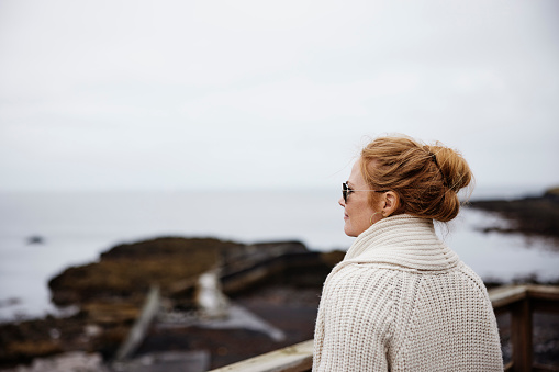 Mature female wearing sweater looking at the seascape. Female tourist on vacation standing on beach cafe deck on an autumn day.