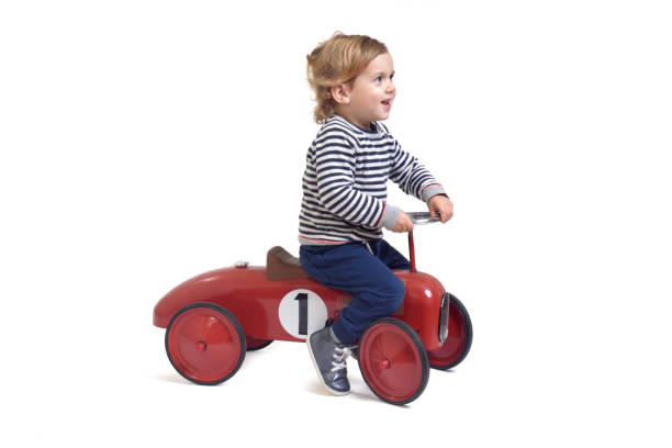 asian young boy is riding on a red toy car, while a baby boy plays with his toy in the living room indoors. This happy child, a little toddler boy, is smiling and having fun driving a big vintage toy car at home