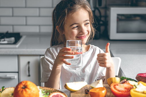Little girl with a glass of water in a kitchen interior with fruits and vegetables.