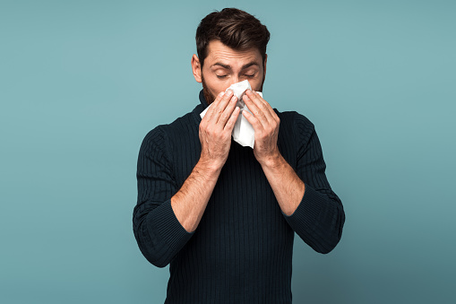 Virus outbreak, flu epidemic. Man sneezing in napkin, cleaning running nose and coughing, suffering influenza symptoms, fever. Indoor studio shot isolated on blue background