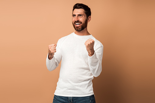 Ecstatic brunette man with beard showing yes i did it gesture, copy space for ad. indoor studio shot isolated on beige background