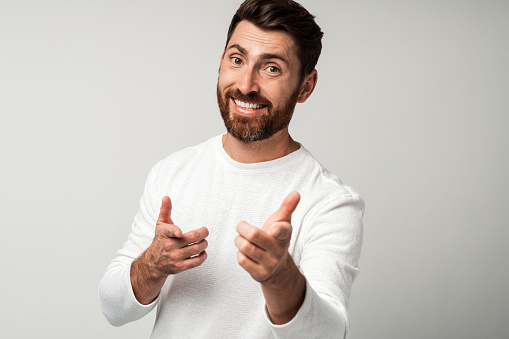 Hey you. Portrait of happy bearded man in white shirt pointing with fingers at somebody against light gray background. Studio shot
