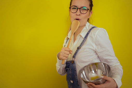 Smiling woman holding a wooden spoon as a microphone and singing and having fun while standing in front of a yellow background