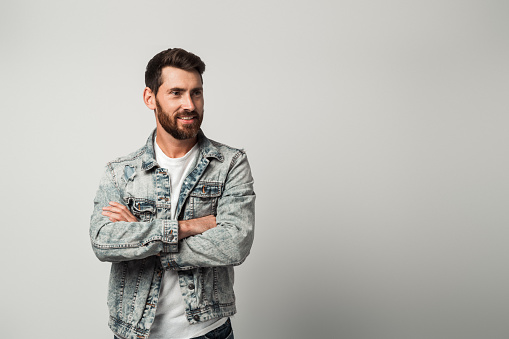 Portrait of bearded handsome man in casual style looking away and smiling while posing arms crossed. Studio shot on grey background