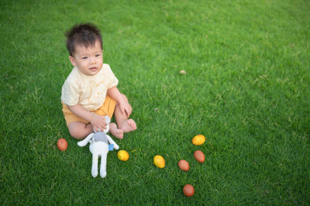 A cheerful one year old boy having a picnic on the lawn, sitting on the grass next to painted colorful Easter eggs during their egg hunt with his favorite bunny toy