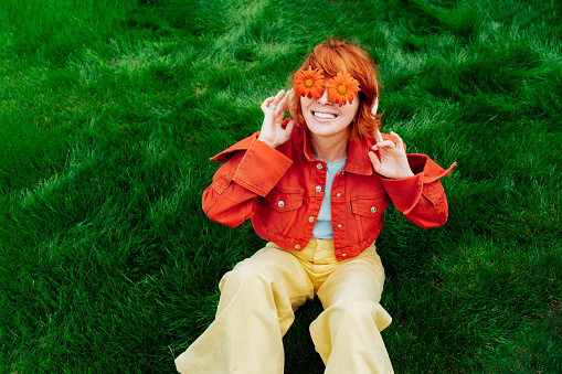 Happy smiling redhead woman with freckles in red orange gerbera Flower glasses sitting on green grass and listening to music in wireless headset. Positive Emotions. Fashion. Spring, summer mood.