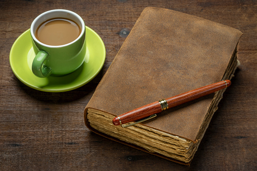 antique leather-bound journal with decked edge handmade paper pages and a stylish pen on a rustic wooden table with coffee, journaling concept