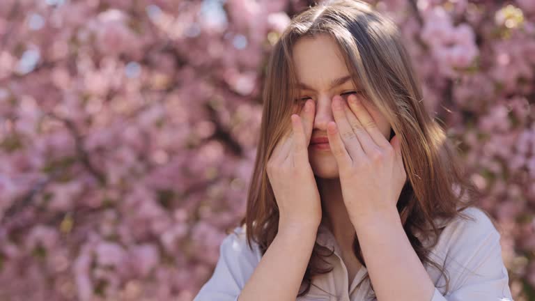 Irritated caucasian young woman standing near blooming sakura tree and scratching itchy eyes during spring time outdoors. Allergic reaction to pollen. For Advertisement of a medicinal product.