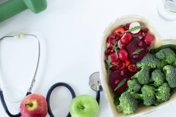 Colorful heart shape from various fruits and vegetables, green dumbbell, and a stethoscope on white, top view stock photo