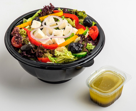 Mixed seasonal salad prepared with chopped cheese, black olive, tomato, capia pepper and lettuce in a black plastic bowl with special dressing on white background. Ready-to-eat.