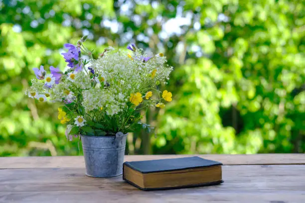 book in black cover, bouquet of wild flowers, family bible lie on wooden table in garden, blurred natural landscape in background with green foliage, concept education, knowledge, christian religion