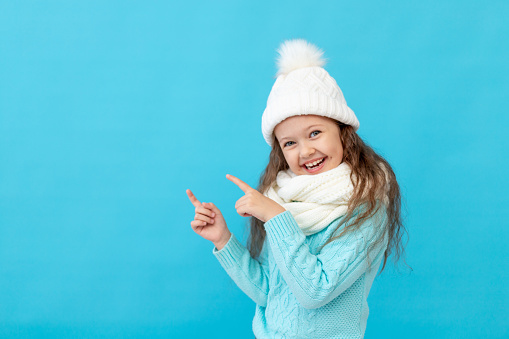 cute little girl child in winter clothes hat and sweater points fingers up at something on a blue isolated background and smiles and laughs, a place or space for text