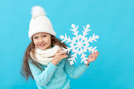 cute little girl child in winter clothes hat and sweater with a big snowflake in her hands on a blue isolated background smiling, a place or space for text