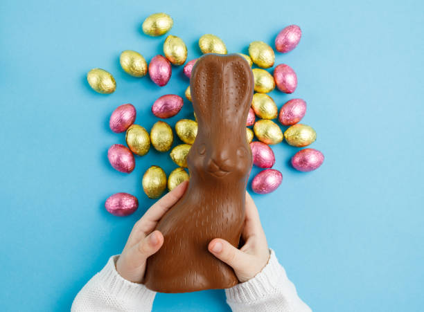 bambino che tiene il coniglietto di cioccolato di pasqua su sfondo blu con le uova di caramelle, concetto di pasqua, vista dall'alto - easter easter egg child chocolate foto e immagini stock