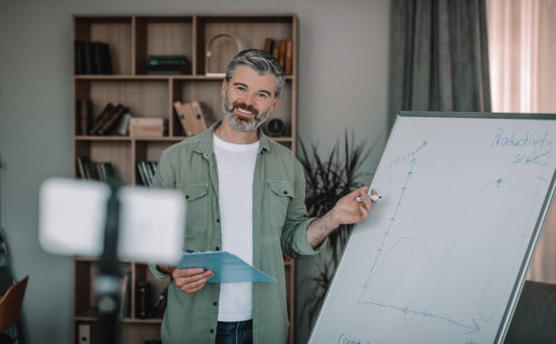 istock Happy elderly european man with beard teacher shooting video lesson on smartphone and points to blackboard 1472383552