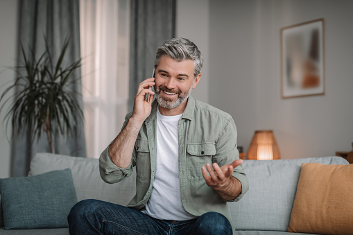 Smiling handsome retired european man with beard calls by smartphone, talk remote in living room interior, copy space. Communication at home alone, conversation in spare time during covid-19 outbreak