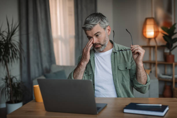 Sad tired retired caucasian man with beard takes off his glasses, rubs eyes looks at computer in room interior stock photo