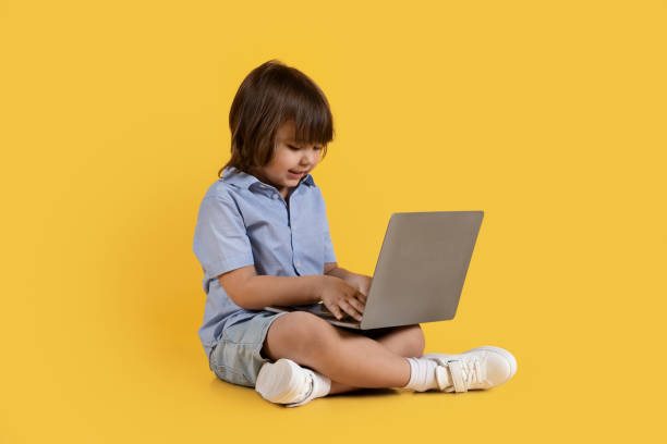 istock Adorable little boy typing on laptop computer, playing developing game, sitting on floor, orange studio background 1472384557