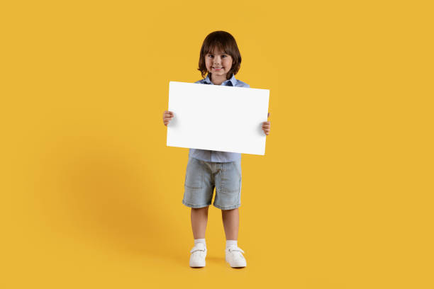 istock Positive little boy holding blank card and smiling to camera, posing over orange studio background, empty space for text 1472384781