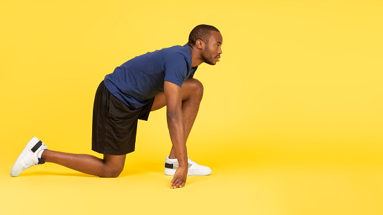 Side View Of African American Runner Guy Standing In Crouch Start Position Looking Aside Over Yellow Background. Male Athlete Posing Ready For Marathon Race In Studio. Sport Of Athletics