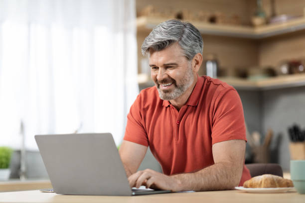 istock Positive mature man typing on laptop keyboard, kitchen interior 1472384957