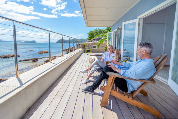 Mature couple drinking wine out on the deck in their waterfront home Mature couple drinking wine out on the deck in their waterfront home. They are happy and smiling sitting in deck chairs. The sea is in the background wealthy house stock pictures, royalty-free photos & images