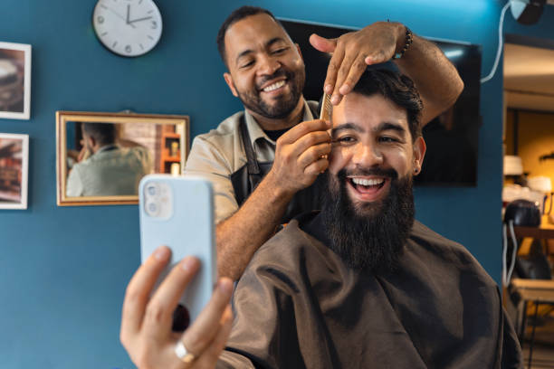 Smiling barber and customer in the barbershop watching a video on a smartphone. Smiling barber and customer in the barbershop watching a video on a smartphone. barber stock pictures, royalty-free photos & images