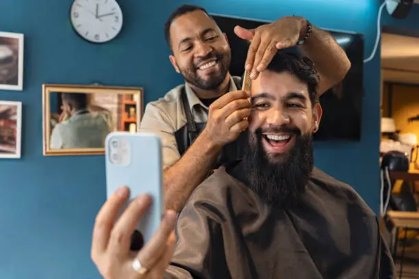 Smiling barber and customer in the barbershop watching a video on a smartphone.