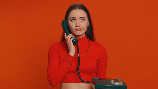Bored, tired pretty young woman in crop top talking on wired vintage telephone of 80s, fooling, making silly faces. Millennial girl having unpleasant conversation isolated on red studio background