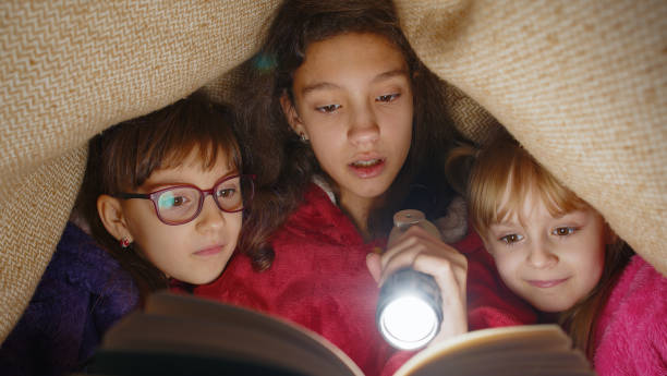 istock Sisters girl kids under blanket reading interesting fairytale story book using flashlight at home 1472397007