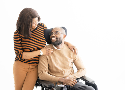 Positive bearded man and woman in casual clothes smiling and looking at each other while sitting in a wheelchair against white background