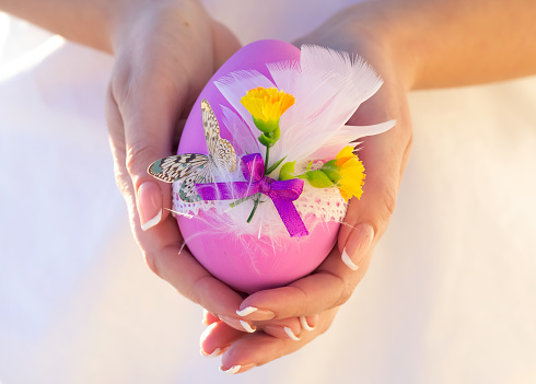 Beautiful crafted pink Easter egg decorated with lace, flowers, feathers and a butterfly in woman's hands with french manicure.