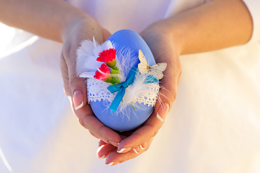 Beautiful crafted blue Easter egg decorated with lace, flowers, feathers and a butterfly in woman's hands with french manicure.