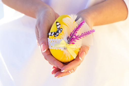 Beautiful crafted yellow Easter egg decorated with lace, flowers, feathers and a butterfly in woman's hands with french manicure.