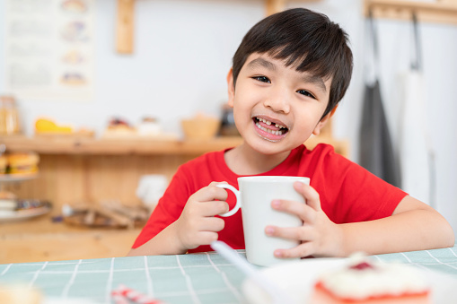 Milk contains all 5 groups of nutrients and develops growth in children. Young Asia boy drink a cup of milk with some cake for the morning meal. Sitting in the kitchen room with smile. Enjoy eating sweet food to increase the energy
