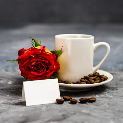 a cup with a rose and wishes on a bright background. Good morning is written on a white sheet, which is next to a rose and a white cup of coffee. Selective focus.