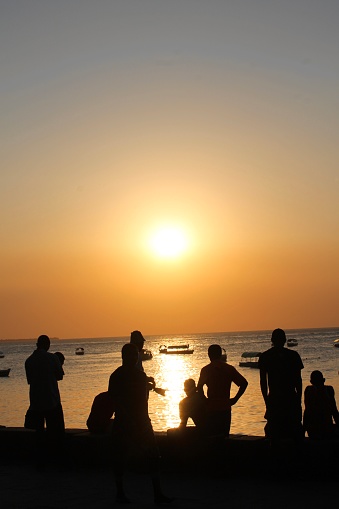 A group of young men watch the vibrant and orange sun set over moored boats from the beach of Stone Town, Zanzibar.