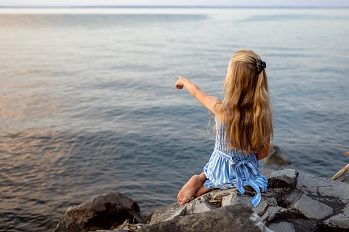Girl with long blond hair sits on a stone on the banks of a river, lake, sea. She is looking at ocean and thinking dreamily. Girl alone outside. Girl sitting on rocks. Vacation on sea.