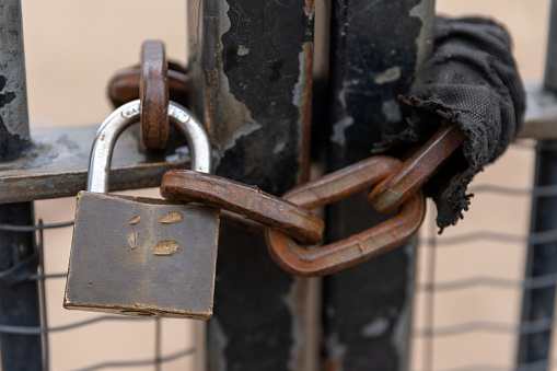 Padlock and chain slung on a gate along a sidewalk. All components are made of metal. Selective focus or shallow depth-of-field image.