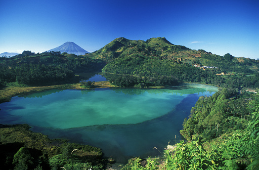Volcanic turquoise color lake, Telaga Warna, on the Dieng Plateau in Central Java, Indonesia. The lake lies in a deep crater with the perfect cone of Mount Sumbing rising in the background. Horizontal color image with clear blue sky and copy space.